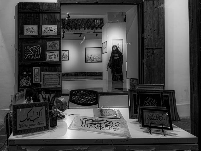 Monochrome interior photograph showing a framed view through a heavy wooden doorway into a gallery. A desk in the foreground holds framed Arabic calligraphy works, art supplies, and a sign in English and Arabic. In the background gallery, framed paintings hang on the walls and a figure stands in profile.