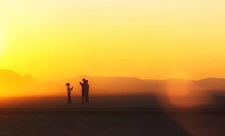 Silhouette of three figures standing against a bright yellow and orange desert sky with distant mountains on the horizon.