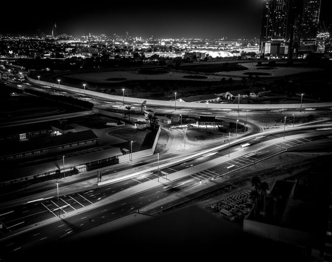 High-angle black and white night shot of urban roads with long-exposure light trails from cars, buildings, and distant city lights.