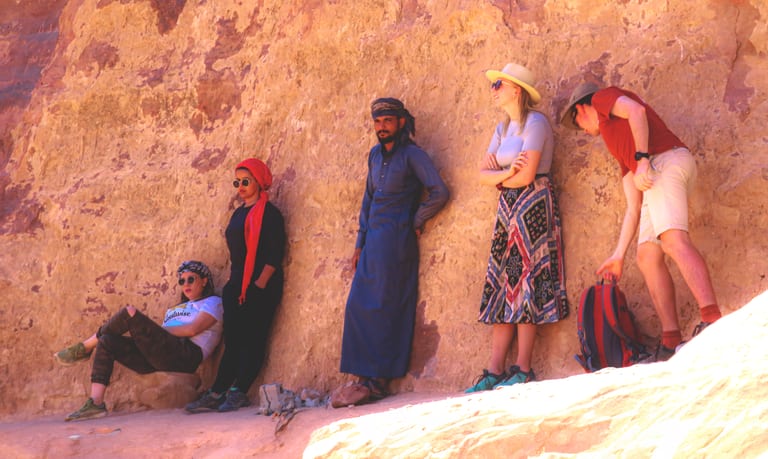 Five people leaning and sitting against a massive, weathered sandstone cliff face, varying in clothing style from casual shirts and skirts to traditional garments and headwraps.