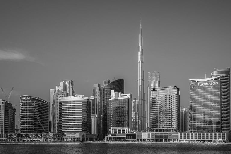 Black and white photograph of a tall skyscraper rising sharply above a crowded city skyline, viewed across a wide body of water with rippling surface texture.