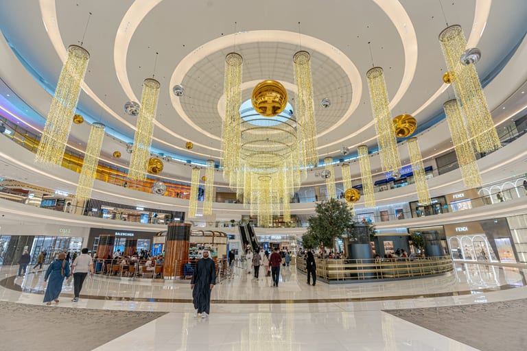 Wide-angle interior view of a grand shopping center atrium with multiple curved balconies, glowing vertical crystal pendant lights, suspended gold spheres, and people walking on polished white marble flooring.