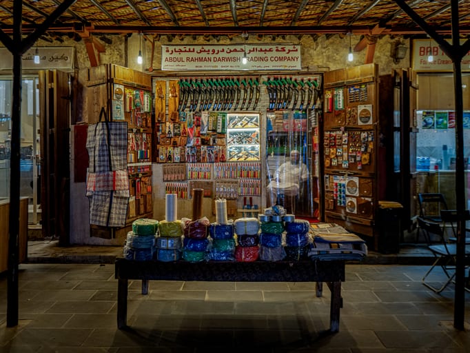 A storefront with an illuminated sign in Arabic and English reads Abdul Rahman Darwish Trading Company, featuring hanging knives, tools, and a seated shopkeeper visible through a clear plastic doorway behind a table of packaged wares.