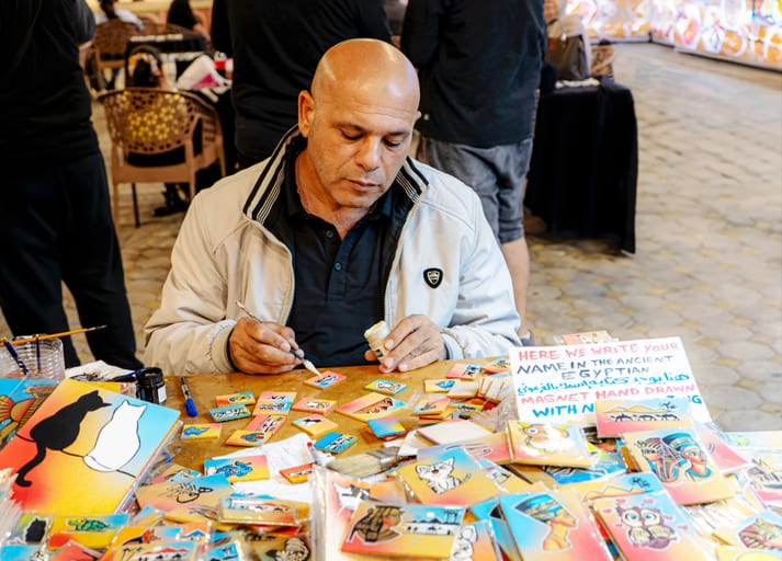 An artisan with a shaved head and light jacket sits at a wooden table covered in small painted tiles and art supplies, holding a fine brush in one hand and a small jar in the other, focused intently on his craft.