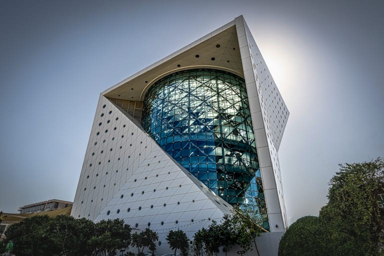 A low-angle architectural shot of a modern building featuring a glass cylinder embedded within a white cubic structure punctured with circular windows, flanked by foliage at the base.