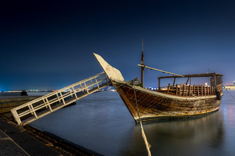 A traditional wooden boat moored at a stone pier at night, connected by a brightly lit white wooden gangway with distant city lights glowing on the horizon.