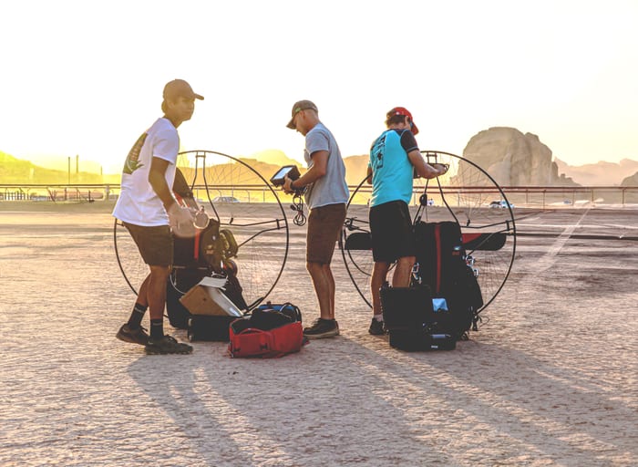 Three people stand outdoors on an open dusty plain in bright backlight, tending to large circular paramotor frames and gear packs on the ground.