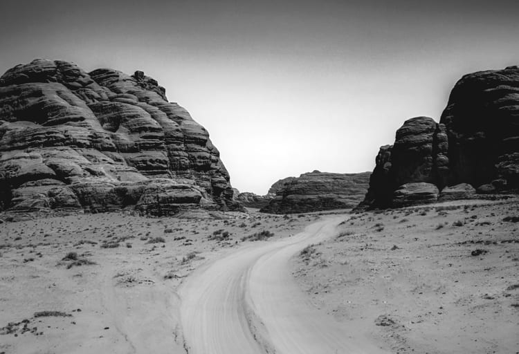 Monochrome landscape photograph of a winding sandy track leading between large, textured rock formations under a bright sky.