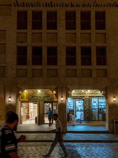 Night street scene showing pedestrians walking in front of a historic multi-story building with symmetrical wooden shutter windows and glowing storefronts at street level.