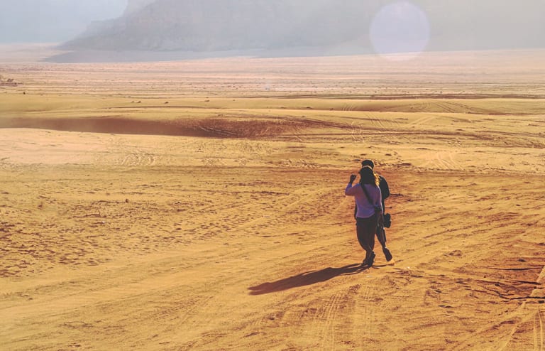 Two people walking away across a vast sandy desert landscape under bright sunlight, casting long dark shadows toward the foreground.