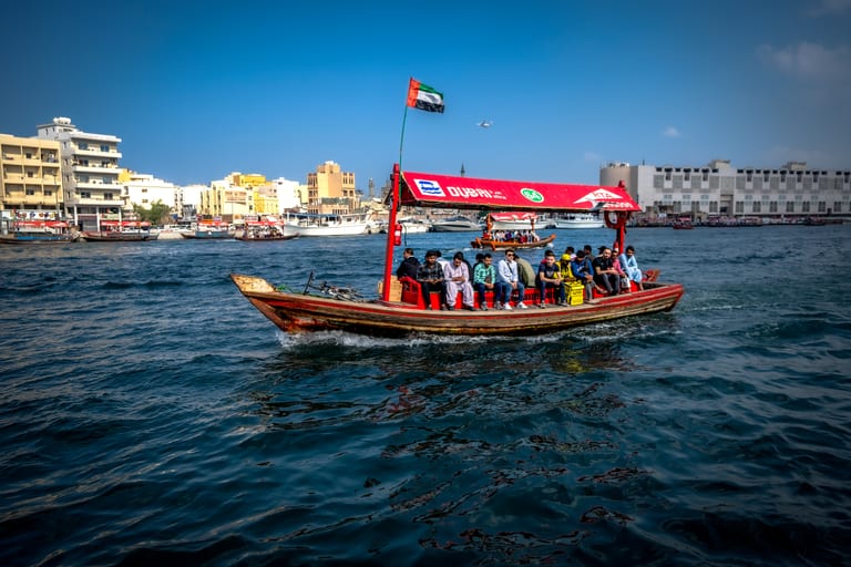 A traditional motorized wooden abra with a red canopy carrying multiple passengers across blue water, with waterfront buildings in the background and an airplane in the sky above.
