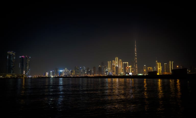 Distant illuminated skyscrapers and a prominent spire at night, viewed across dark water reflecting the city lights.