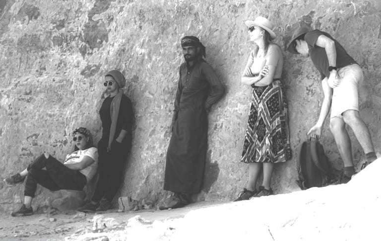 A black-and-white horizontal photograph showing a line of five diverse individuals resting and leaning against a rough rock wall, captured from left to right in varying postures from seated to standing.
