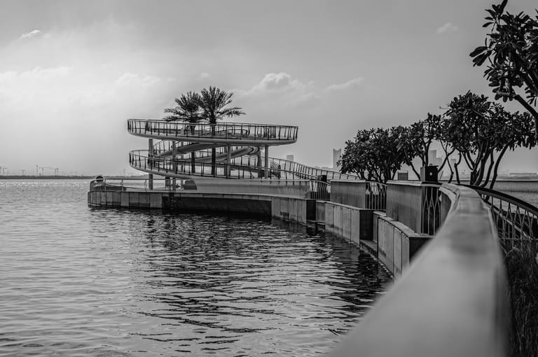 Black and white photograph of a curved waterfront walkway and a multi-level spiral viewing platform with palm trees, alongside rippling water and distant hazy horizons.