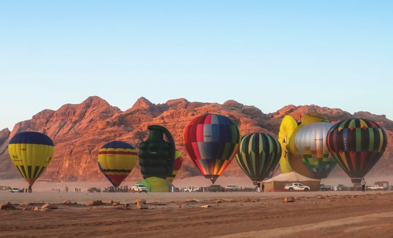 Wide shot of several large hot air balloons in various vibrant patterns lined up on the desert floor beneath a range of rugged sandstone mountains.