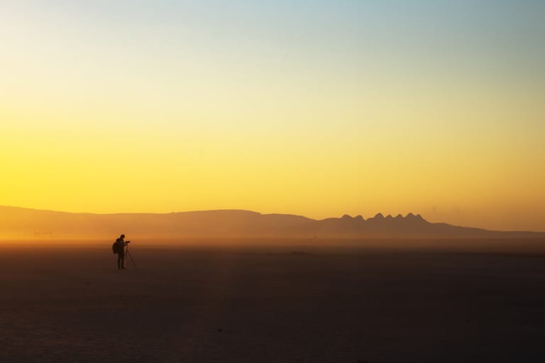 Wide-angle landscape shot of a silhouetted photographer with a tripod in a flat desert beneath a bright yellow and blue twilight sky, with distant mountains along the horizon.