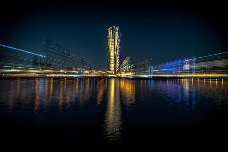 Night long-exposure photograph showing streaking golden and blue city lights radiating outward from a central illuminated skyscraper, reflecting onto dark water below.