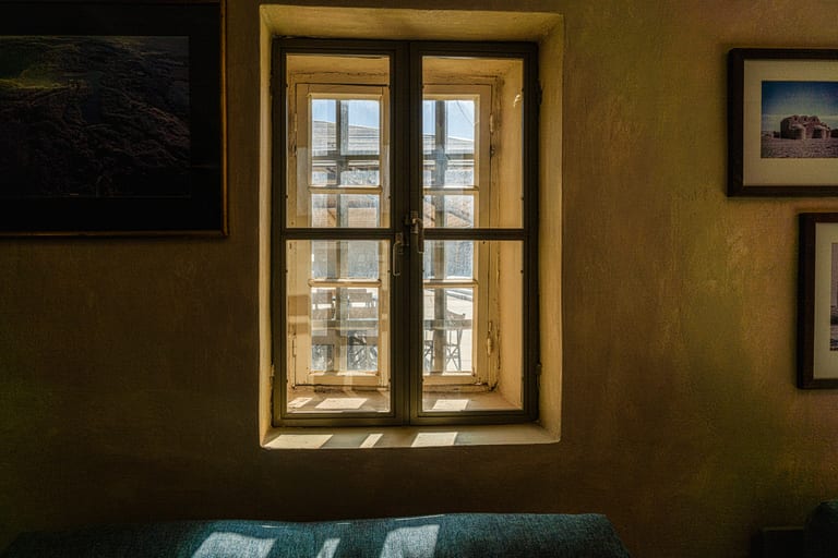 A centered double-paned window set in a textured beige interior wall, flanked by framed photographs, with sunlight casting sharp shadows on the sill and a blue upholstered seat in the foreground.
