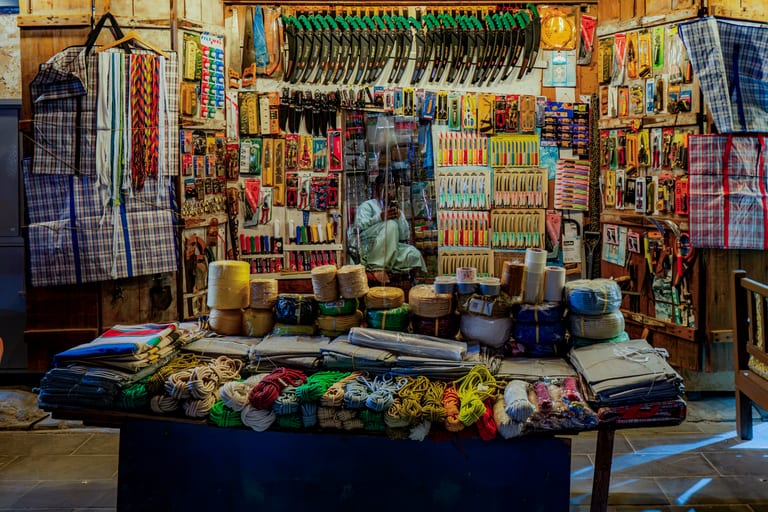 Interior view of a small shop featuring wooden walls completely covered in hanging knives, saws, and tools, with spools of rope and textiles neatly stacked on a wooden counter in the foreground and a person seated in the background.