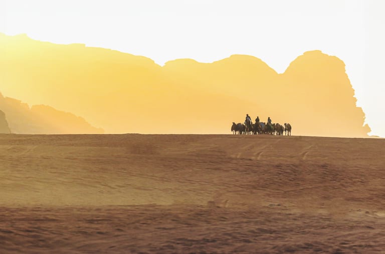 A group of people riding camels in a silhouetted caravan across a golden desert dune under bright backlit sky, with large mountain formations in the background.