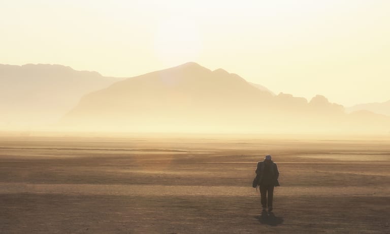 A lone figure walking across a vast, dusty plain towards distant hazy mountains under a bright, diffused sun.