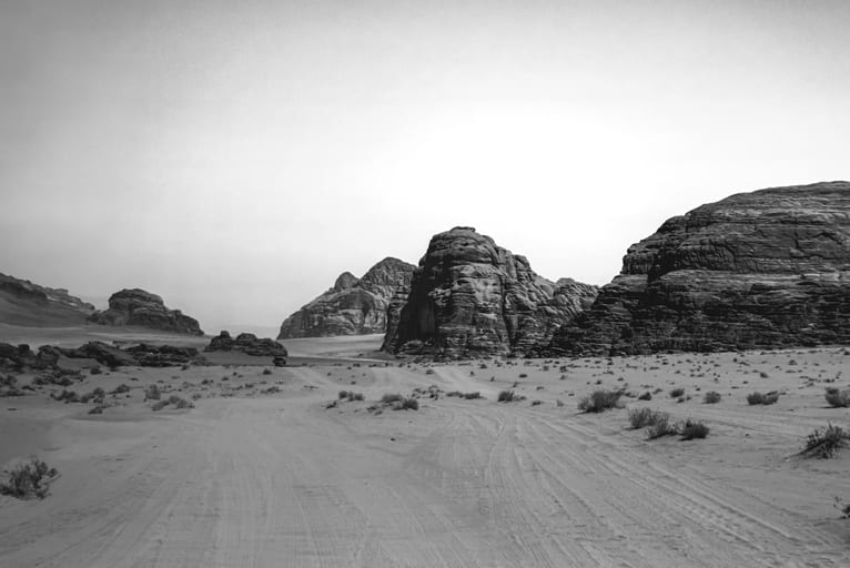 Monochrome landscape view of rugged sandstone rock formations rising from a desert floor with vehicle tracks and scattered desert shrubs in the foreground under a pale sky.