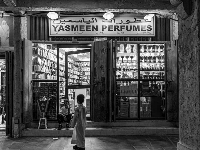 Black and white photograph of a storefront named Yasmeen Perfumes with two children standing and sitting on the stone step outside, flanked by displayed glass bottles and merchandise.