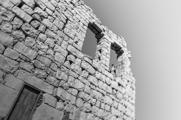 Low-angle monochrome photograph of an ancient stone wall with rough-hewn blocks, two upper window cutouts against a bright sky, and a closed dark door at the lower left.