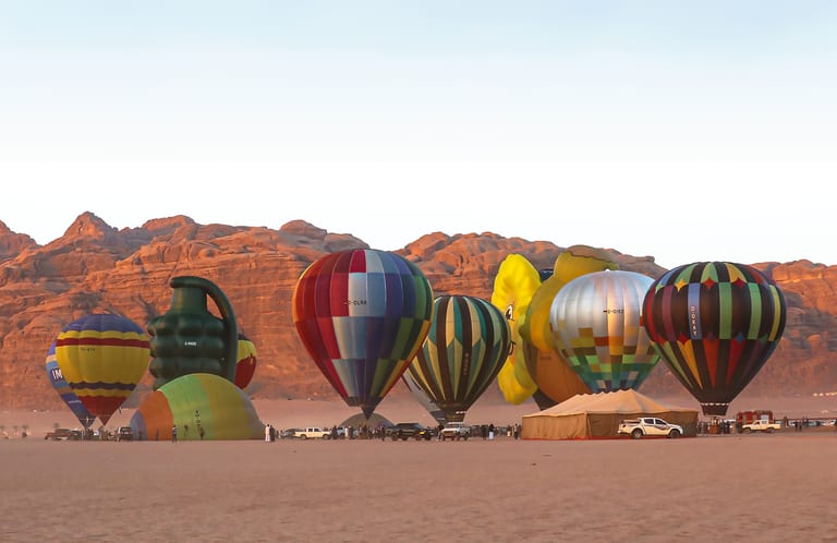 Wide shot of multiple hot air balloons in various colors and unusual shapes standing or partially inflated on a vast desert floor against a backdrop of rocky mountains under a clear sky.