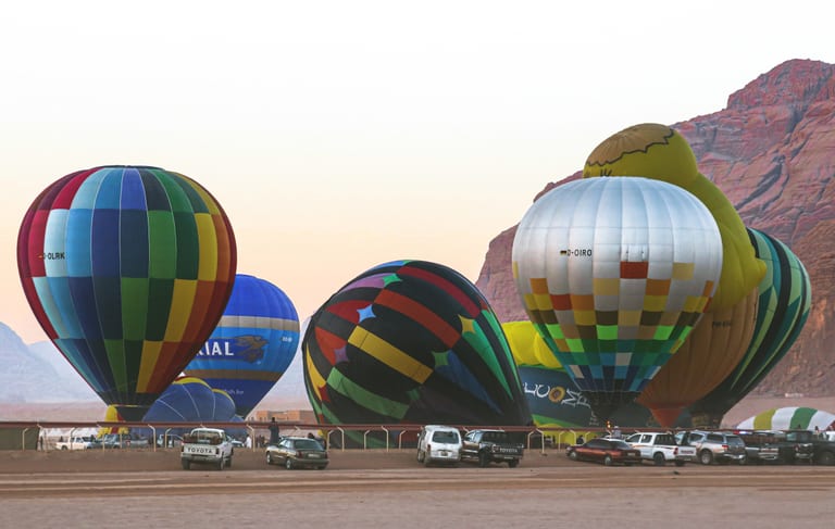 A line of colorful hot air balloons stands inflated on flat desert ground in front of rocky sandstone mountains under a clear sky, with parked pickup trucks visible in the foreground.
