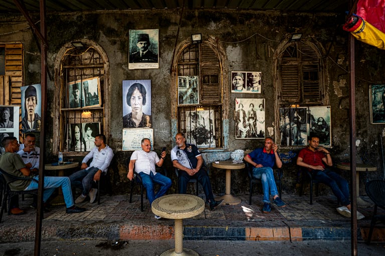 Horizontal color photograph showing six men sitting along a textured stone wall lined with old portraits and arched shuttered windows, separated by small plastic tables on a paved sidewalk.
