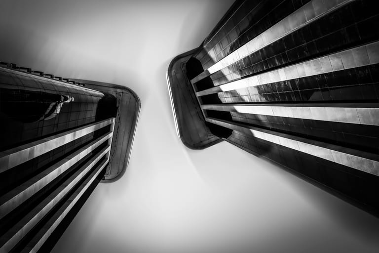 A low-angle monochrome shot looking up at the facades of two tall modern buildings against a bright, smooth sky, creating sharp diagonal lines and high-contrast geometric patterns.