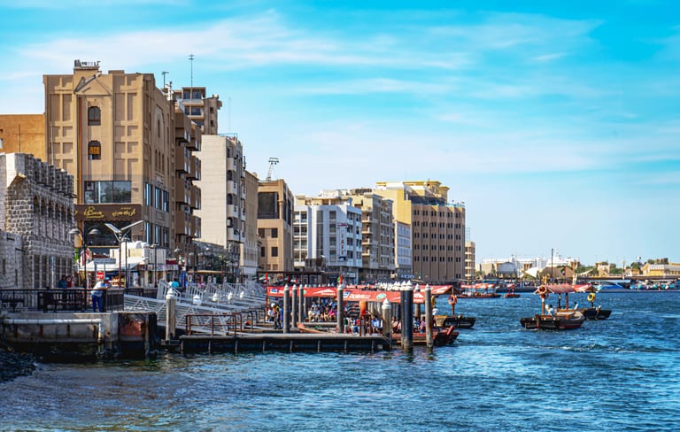 Daytime exterior view of wooden boats docked at a pier beside a busy waterfront promenade, with multi-story buildings lining the shore under a bright blue sky.