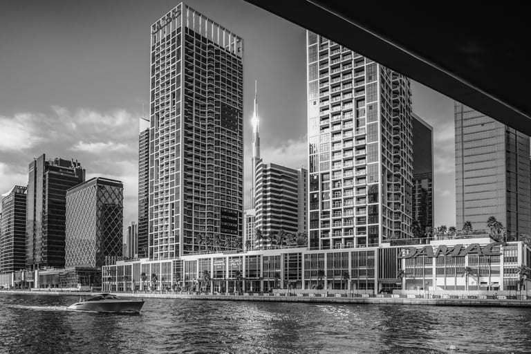 Monochrome eye-level photograph of a modern city canal with a sleek motorboat moving across the water in the lower left. In the background, tall skyscrapers and a distant slender tower rise beneath a bright sky, framed by an overhead architectural canopy in the upper right.