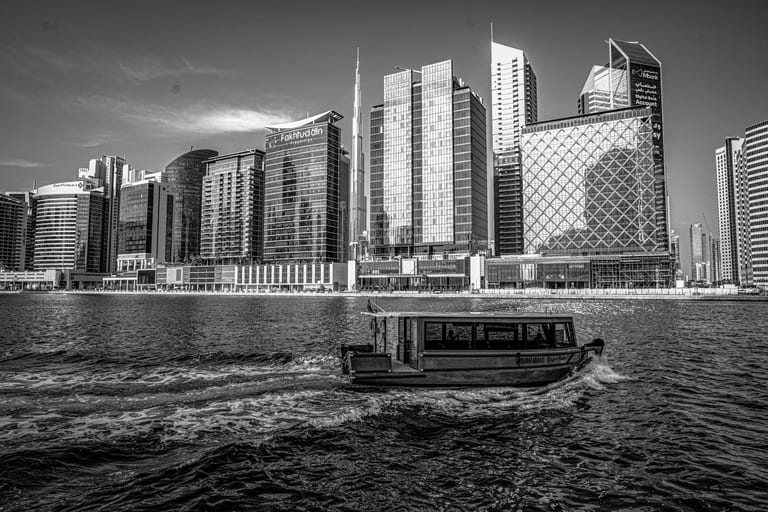 A black-and-white photograph of a passenger boat cruising across dark water from right to left, leaving a foamy white wake, with a skyline of tall contemporary buildings in the background.