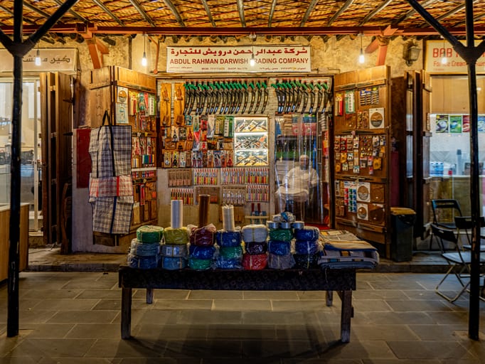 A store facade displaying rows of saws and tools with a seated figure inside, and a wooden table piled with wrapped bundles of goods in the foreground.