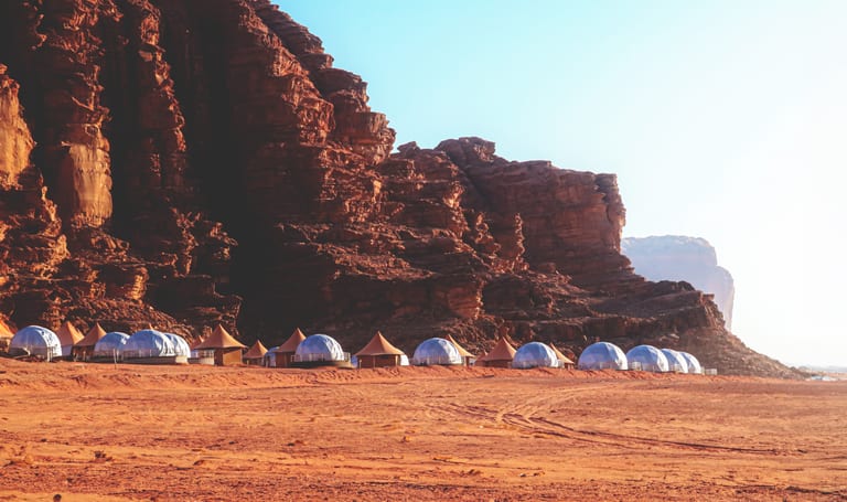 A wide-angle landscape shot showing a line of white dome-shaped tents and brown conical structures nestled at the base of massive, textured red sandstone cliffs under a clear bright sky.