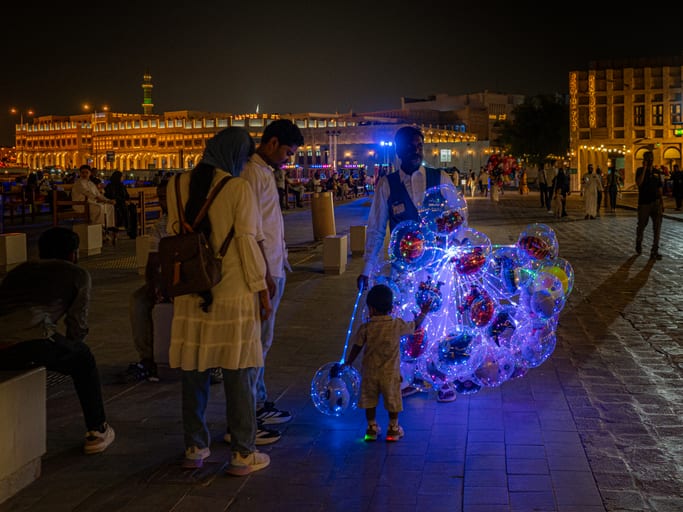 A street scene at night where a young child reaches up to touch a cluster of bright blue glowing LED balloons held by a vendor while two adults stand watching on a stone promenade.