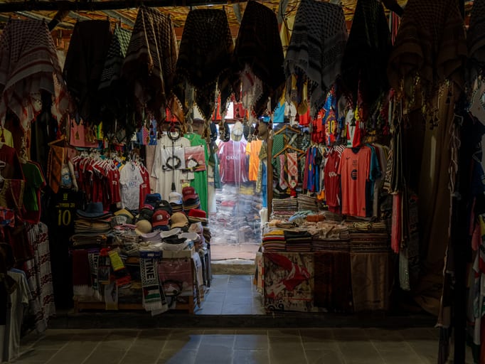 A market stall laden with hanging patterned headscarves, folded textiles, and sports jerseys, viewed from a stone-paved floor looking inward toward a bright open doorway.