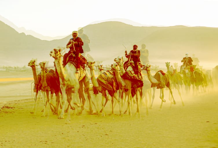 A group of riders on camels moving across a sandy landscape in a line, rendered in warm golden tones with visible motion blur and dust kicked up from the ground.
