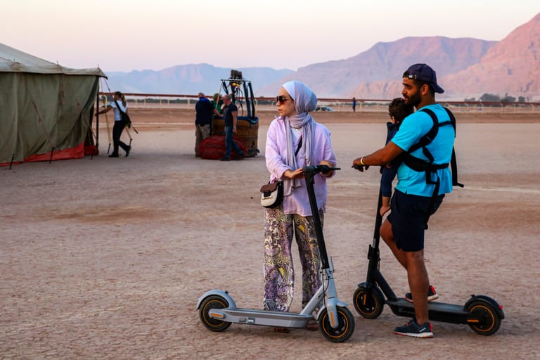 A woman wearing a hijab and patterned trousers and a man carrying a young child in a harness both stand on electric kick scooters on a dusty, dry plain outdoors, with a tent, hot air balloon basket, and distant mountains in the background under soft daylight.