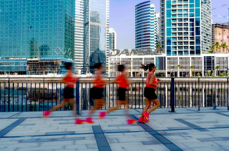 A motion-blurred sequence of a runner wearing bright red shoes and an orange top moving across a tiled urban promenade with towering skyscrapers across a body of water in the background.