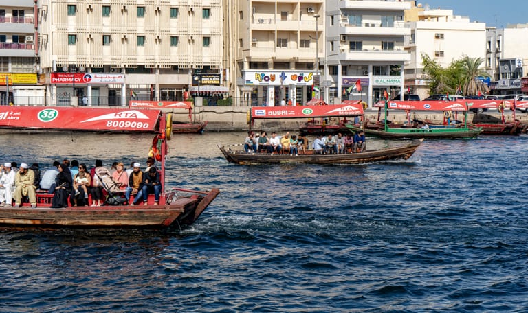 Multiple traditional wooden passenger boats filled with people crossing a blue water channel, with low-rise apartment and retail buildings lining the far bank under bright daylight.