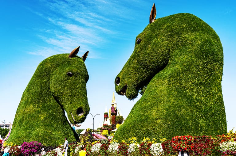 Low-angle shot of two giant green topiary horse heads facing each other under a clear blue sky, with colorful flower beds lining the foreground and a small castle structure visible in the gap between them.