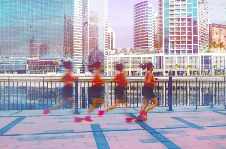 A woman running along a tiled waterfront promenade in Dubai, captured in a multi-exposure technique showing sequential blurred figures leading up to her sharp final stride, with modern skyscrapers in the background.
