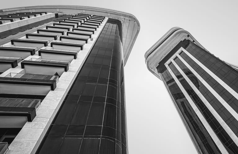 A low-angle black-and-white photograph of two tall modern high-rise buildings, showing dark glass panels, rows of balconies, and curved architectural lines against a plain sky.