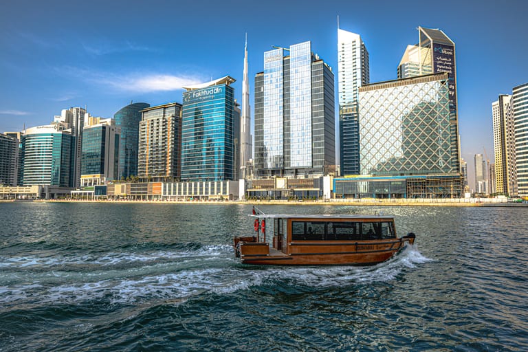 A brown motorized water taxi moving across textured blue water in the foreground, with a line of tall modern skyscrapers and a slender spire rising against a blue sky in the background.