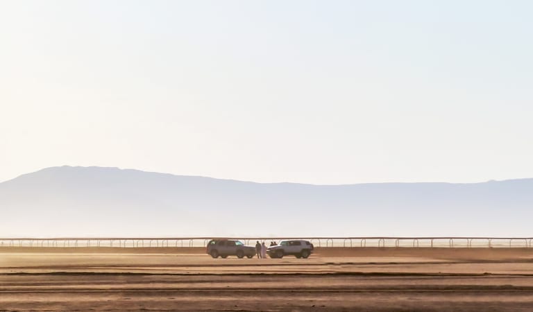 Wide-angle view of a vast desert expanse with two parked SUVs and several small human figures standing between them near a long horizontal fence, backed by faint hazy mountains and a pale sky.