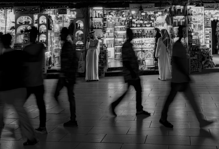 Monochrome street scene showing several blurred figures walking across a tiled walkway in the foreground, with a sharp, stationary man in a traditional thobe standing outside a brightly illuminated shop entrance in the background.