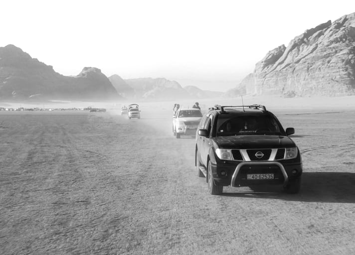Monochrome photograph of multiple off-road vehicles driving in a procession across a dusty desert terrain toward distant rocky hills.